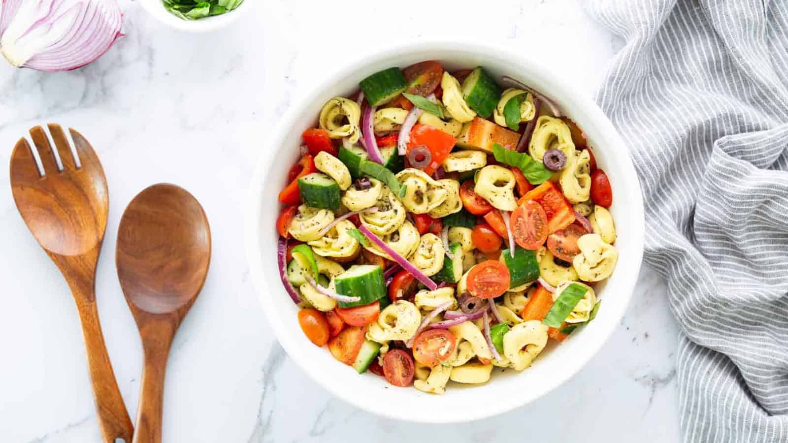 Bowl of tortellini pasta salad with veggies on a marble surface, next to wooden utensils and a striped towel.