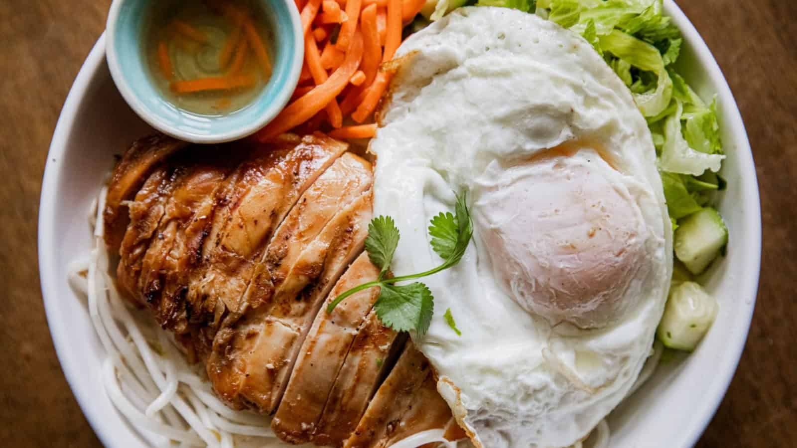 Bowl of noodles topped with grilled chicken, a fried egg, pickled carrots, lettuce, cucumber, and dipping sauce.