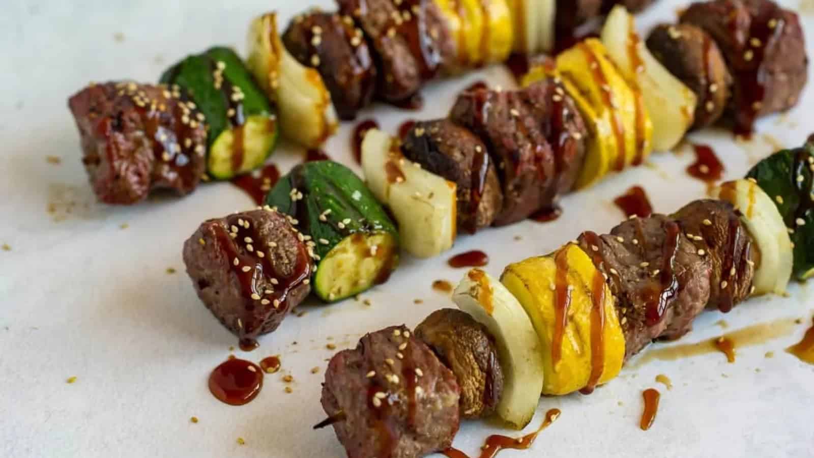 Grilled beef and vegetable skewers with zucchini, squash, onion, and sauce drizzled on parchment paper.