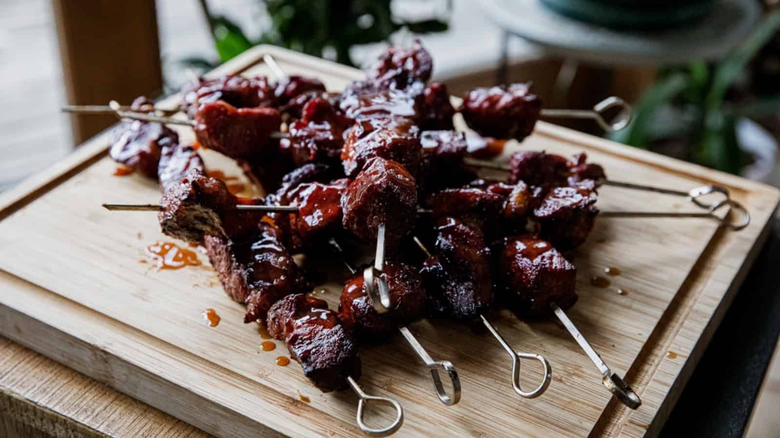 Barbecue meat skewers stacked on a wooden board, glistening with sauce, ready to be served.