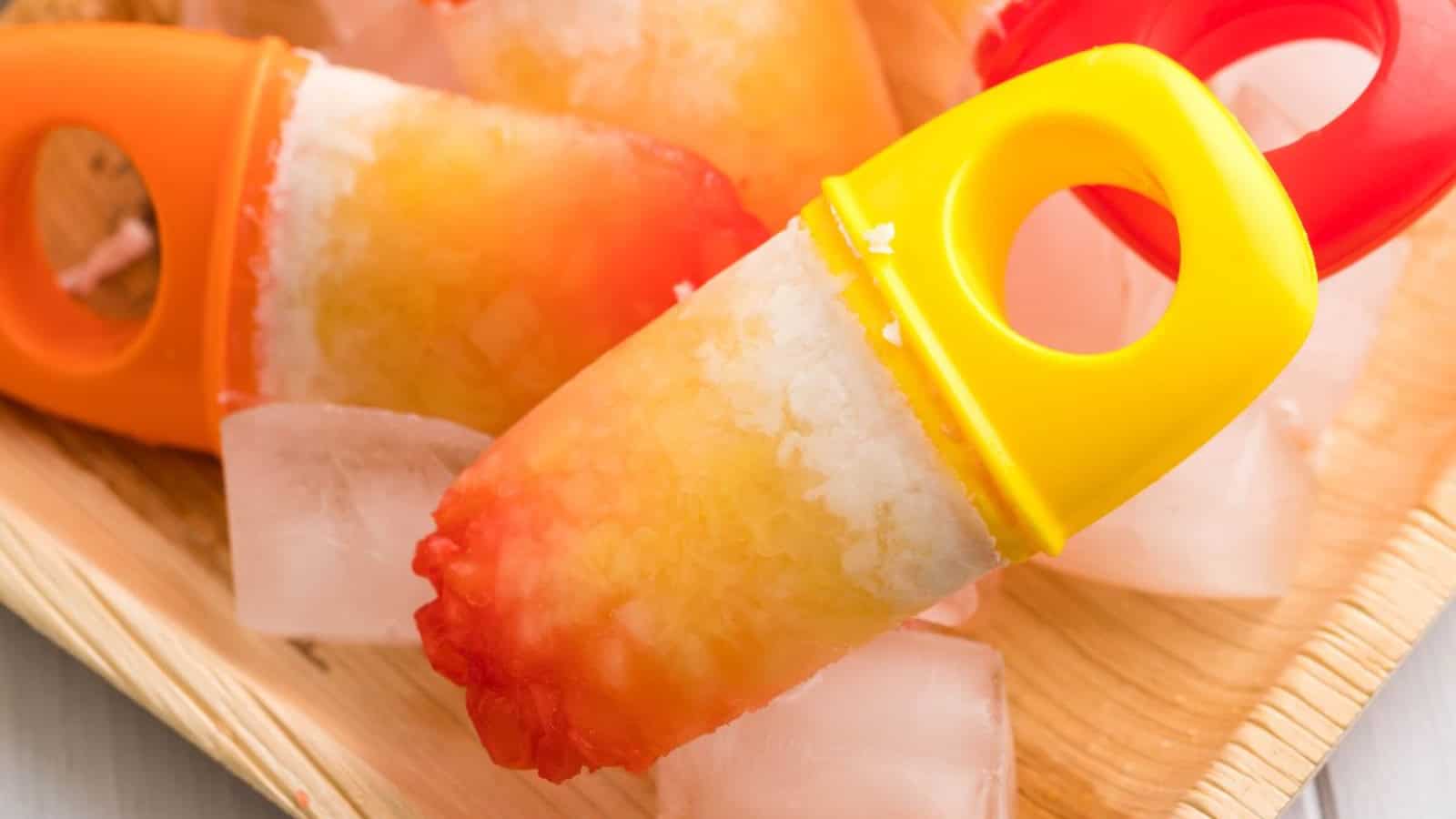Colorful homemade popsicles with ice cubes on a wooden tray, featuring yellow and orange shades.