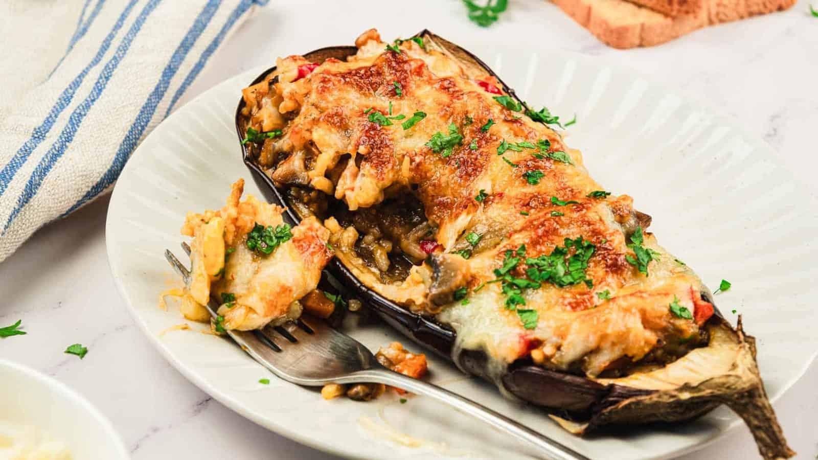 Baked stuffed eggplant topped with melted cheese and herbs on a white plate, with a fork beside it.