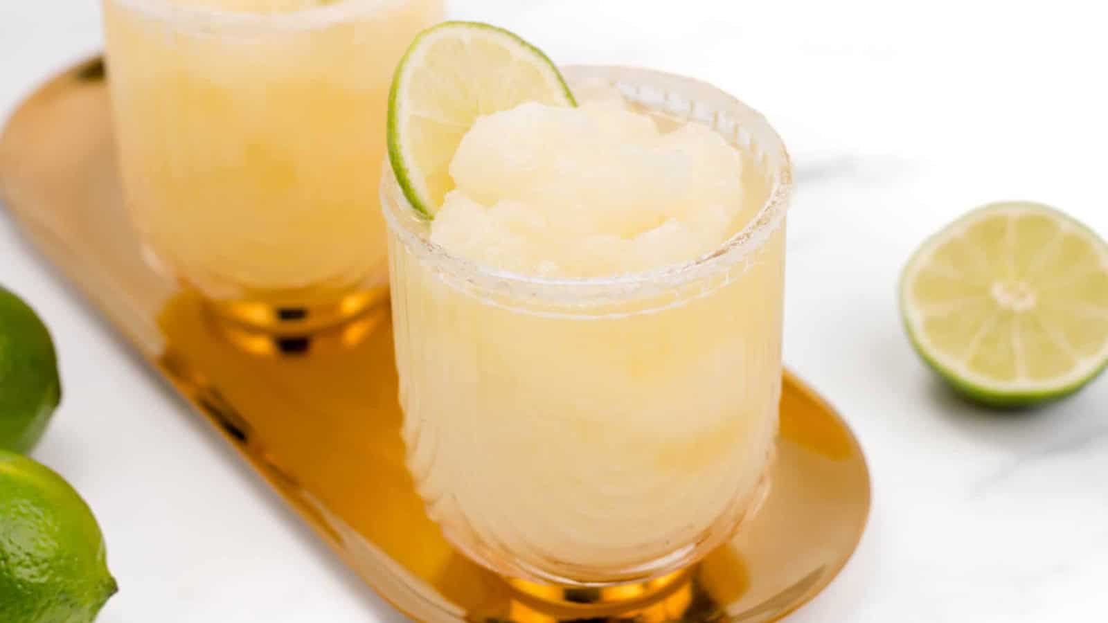 A frozen margarita in a glass garnished with a lime slice, next to fresh limes on a gold tray.