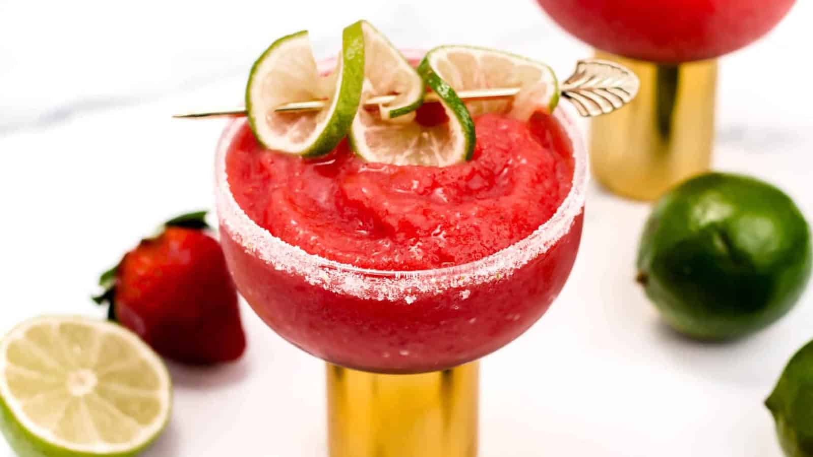 A strawberry margarita in a salted glass, garnished with lime slices, with limes and a strawberry nearby.