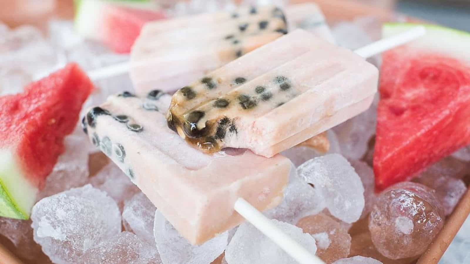 Two pink popsicles with black seeds rest on ice beside slices of watermelon.