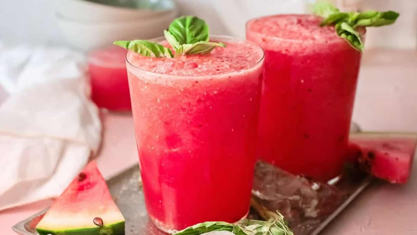 Two glasses of bright pink watermelon smoothie garnished with basil leaves, with watermelon slices nearby.