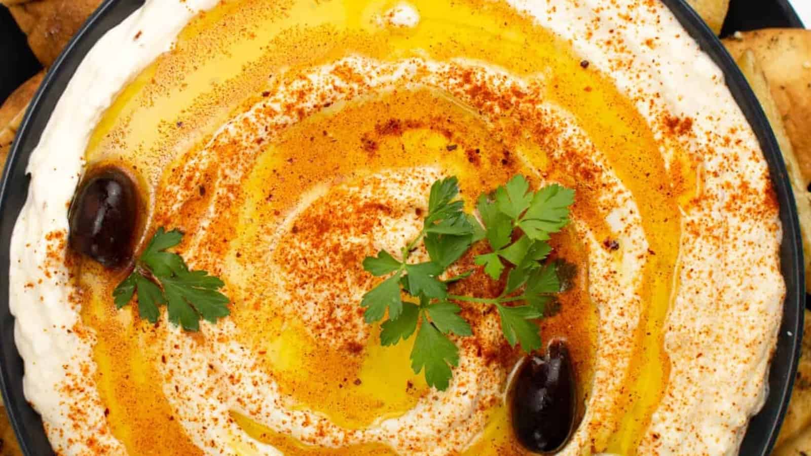 A bowl of hummus topped with olive oil, paprika, parsley, and two olives.