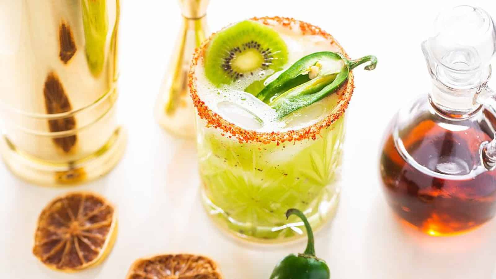 A green cocktail with kiwi, jalapeño, and chili rim, beside syrup, shaker, and dried lime slices.