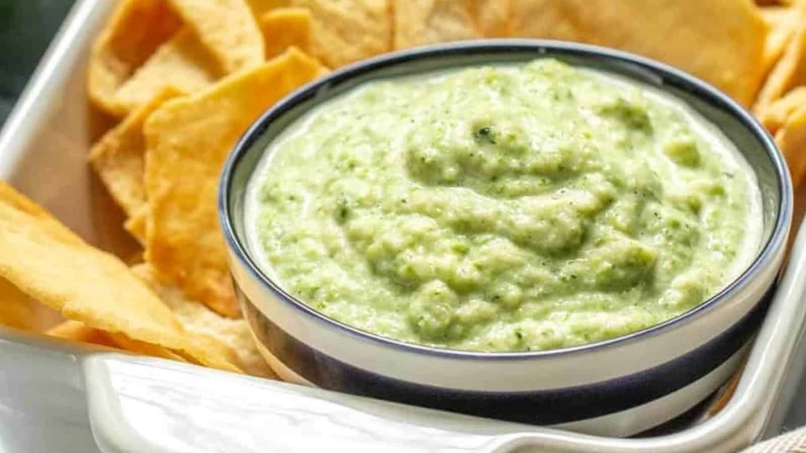 Bowl of creamy green dip surrounded by crispy pita chips on a white tray.