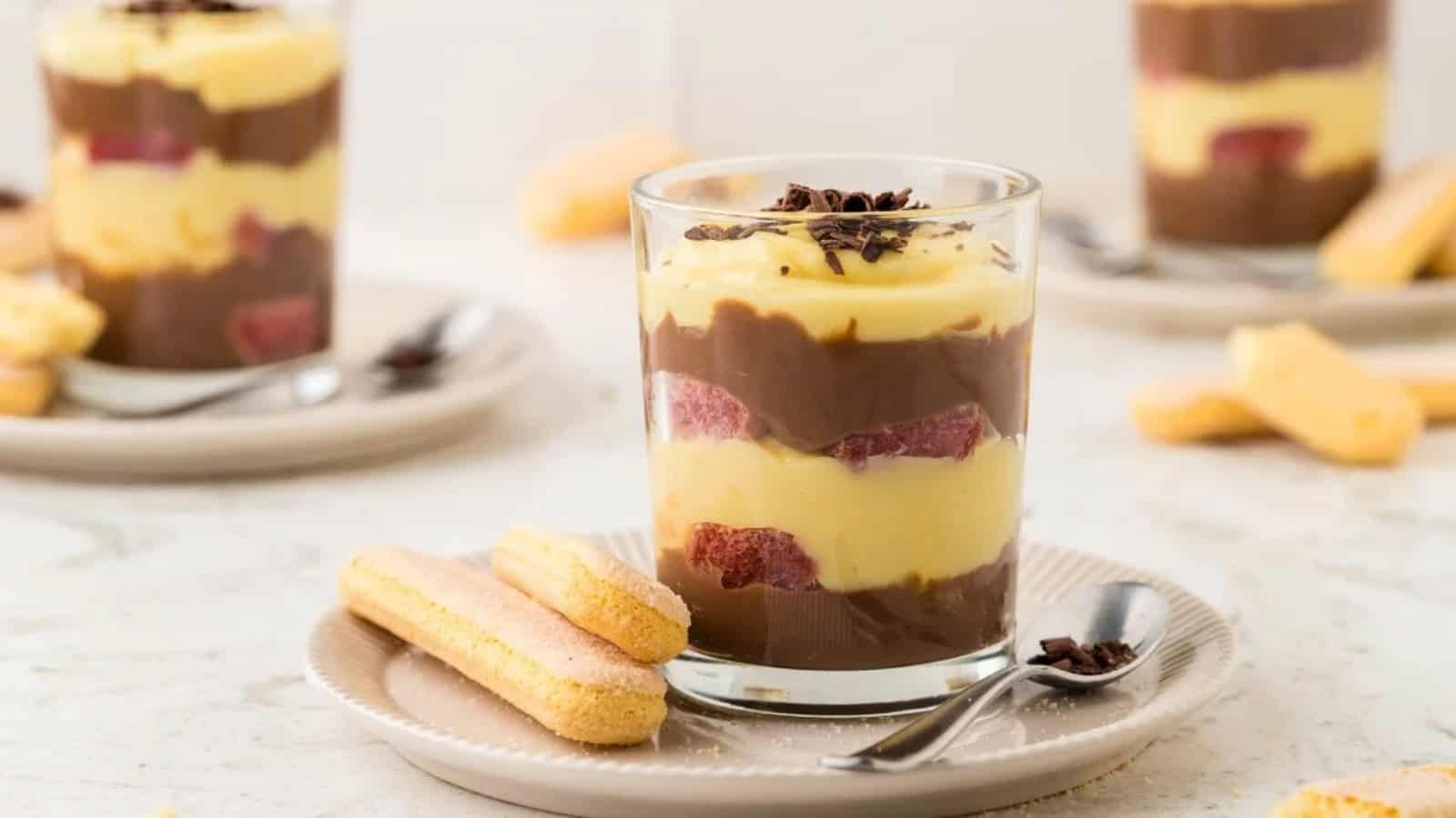 A glass dessert with chocolate and vanilla layers, strawberries, and ladyfingers on a plate with a spoon.